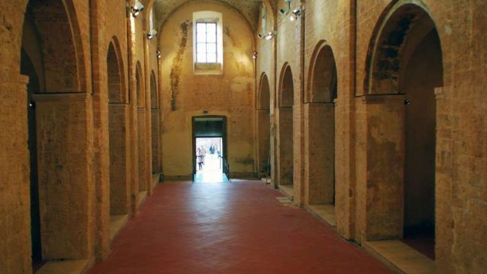 Interno di una sala in mattoni con navata centrale e serie di arcate laterali su colonne, pavimento in cotto rosso e soffitto a volta. In fondo si vedono una finestra alta e una porta aperta che dà verso l’esterno, con illuminazione a faretti lungo le pareti.