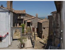 Vista di un piccolo borgo con case in pietra e tetti in tegole, cortili e scalinate in salita tra vicoli stretti. Un arco in muratura e muri intonacati incorniciano la scena, con colline sullo sfondo sotto un cielo sereno.