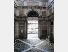 Interno di un cortile in pietra con pavimentazione a motivi geometrici e pareti con riquadri in mattoni, centrato su un grande arco con cancello in ferro battuto aperto. Oltre l’arco si vede una balaustra in pietra e la facciata di un edificio esterno.