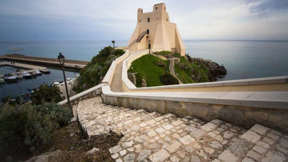 Sentiero in pietra in discesa che conduce a una torre-fortezza chiara su un promontorio, con mura e scalinate affacciate sul mare. Sul lato sinistro si vede un piccolo porto con pontile e barche, circondato da vegetazione costiera.