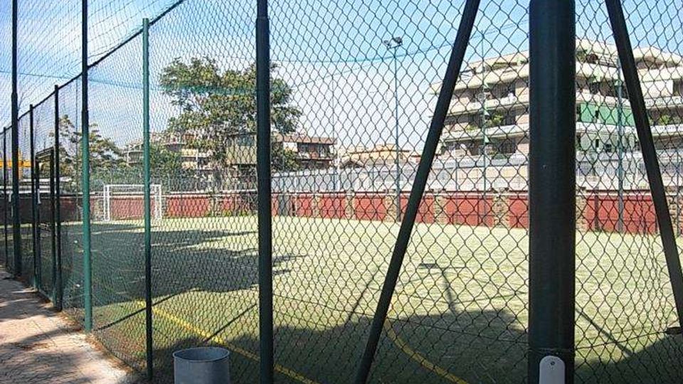 Campo sportivo all’aperto con superficie in erba sintetica e linee gialle, circondato da alte recinzioni in rete metallica e illuminazione su pali. In primo piano si vede un marciapiede in pietra lungo la recinzione, con edifici residenziali sullo sfondo.
