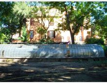 Serra bassa e allungata con struttura metallica e pannelli traslucidi, situata in un’area ombreggiata con terreno scoperto. Sullo sfondo si vede un edificio a più piani color ocra con persiane, circondato da alberi e vegetazione.