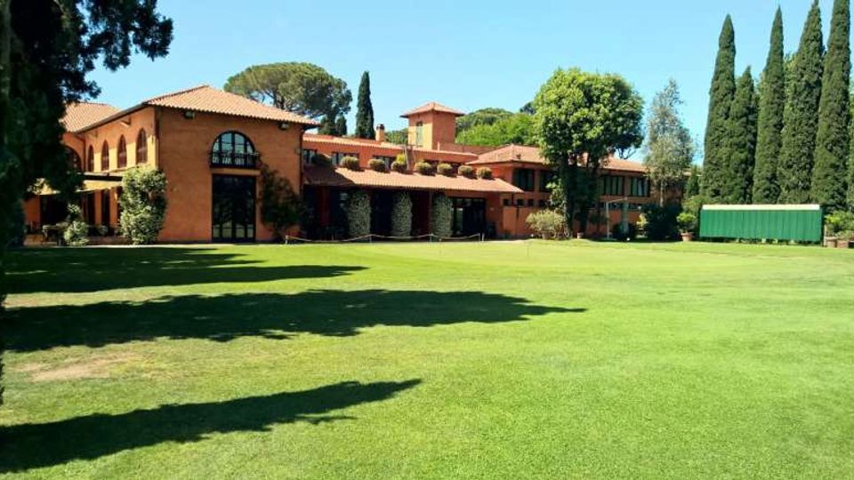 Grande edificio in mattoni con tetti in tegole rosse e ampie finestre ad arco, affacciato su un vasto prato verde. Intorno ci sono alti cipressi e altri alberi, con una struttura verde rettangolare sul lato destro.