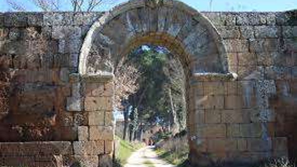 Antico arco in pietra con apertura a tutto sesto inserito in un tratto di mura in blocchi, con segni di degrado e vegetazione. Attraverso l’arco si vede un sentiero che prosegue in un’area alberata sotto un cielo limpido.