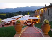 Terrazza all’aperto con tavoli e sedie sotto grandi ombrelloni bianchi, accanto a un edificio in pietra illuminato e raggiungibile tramite una scalinata in cotto. Sullo sfondo si vedono colline e montagne, con grandi vasi decorativi ai lati delle scale e aree verdi intorno.