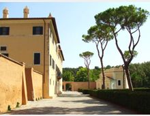 Viale con pavimentazione in ghiaia accanto a un grande edificio storico color ocra con persiane scure e tetto in coppi. Sul lato opposto ci sono siepi potate e alti pini marittimi, con un piccolo edificio e un arco d’ingresso sullo sfondo.