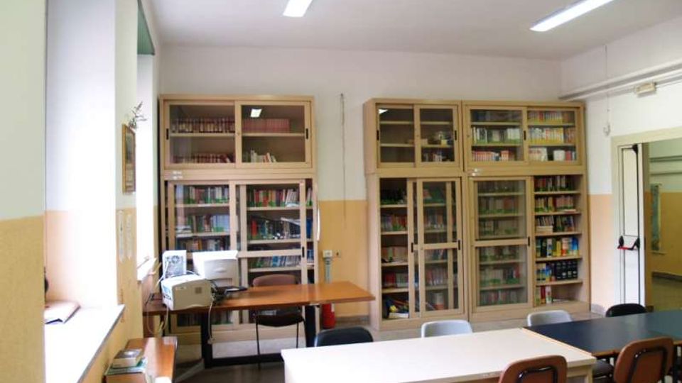 Aula adibita a biblioteca con armadi vetrati pieni di libri lungo le pareti e tavoli rettangolari con sedie in primo piano. Sul lato sinistro ci sono finestre e un banco con un computer, mentre il soffitto è illuminato da lampade fluorescenti.