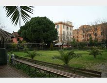 Giardino urbano con prato e piccole palme, una lunga panchina in legno in primo piano e un grande albero sempreverde al centro. Sullo sfondo si vedono edifici residenziali di più piani e alberi spogli lungo il perimetro.