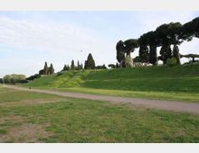 Ampio parco con prato in pendenza e un sentiero sterrato che attraversa l’area. Sullo sfondo si vedono alberi alti e sagomati, tra cui cipressi e pini, lungo il crinale erboso.