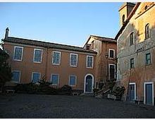 Cortile lastricato con panchine e alcune piante, circondato da edifici in muratura color ocra e mattoni con finestre regolari. Sullo sfondo si vedono porte ad arco e facciate storiche illuminate dalla luce bassa, sotto un cielo sereno.