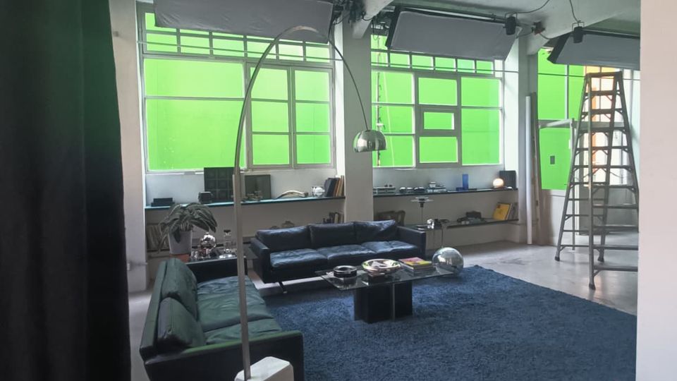 Interno di un ampio loft con soffitto in cemento a vista e grandi finestre a riquadri schermate da pannelli verdi. Area soggiorno con divani, tavolino basso su tappeto blu, lampada ad arco e una scala a pioli vicino alle finestre.