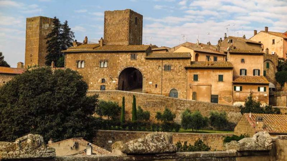 Borgo medievale in pietra su una collina, con edifici dai tetti in tegole e una grande porta ad arco nelle mura. Sullo sfondo si vedono torri quadrate e case addossate, con alberi e vegetazione ai piedi delle fortificazioni.