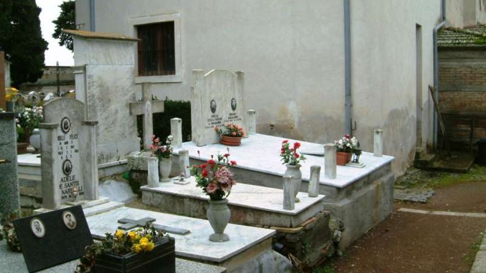 Piccolo cimitero con tombe in marmo e lapidi con croci e vasi di fiori, disposte su piattaforme rialzate. Sullo sfondo si vede il muro esterno chiaro di un edificio e un vialetto laterale in cemento.