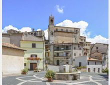 Piazza di un borgo storico con pavimentazione in pietra a motivi curvi e una fontana centrale. Sullo sfondo si vedono case in muratura e un campanile in pietra che svetta tra edifici con tetti in coppi, sotto un cielo azzurro con nuvole.