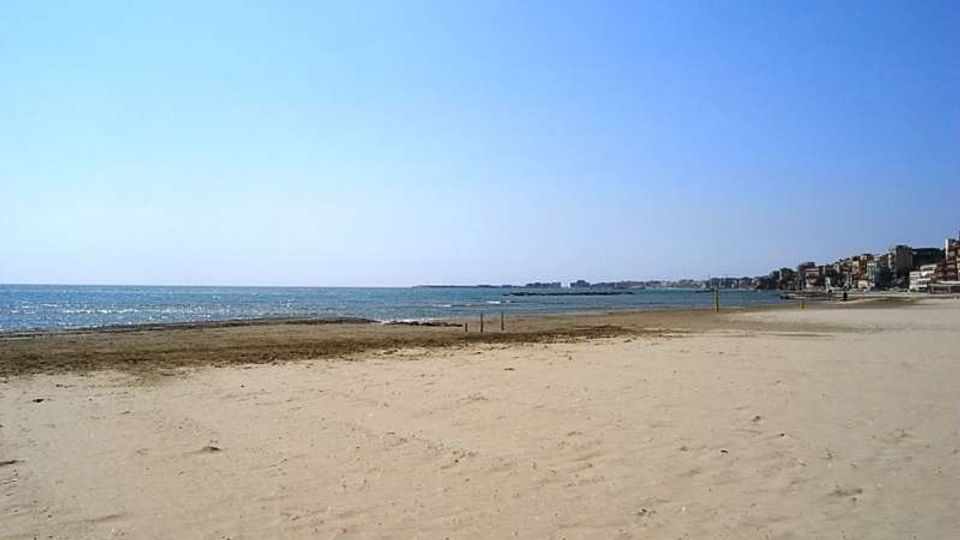 Ampia spiaggia sabbiosa sul mare con linea di battigia e acqua calma sotto un cielo sereno. Sullo sfondo, a destra, si vede la costa con edifici lungo il litorale e un promontorio all’orizzonte.