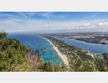 Veduta panoramica dall’alto di un lungo litorale sabbioso con mare turchese, affiancato da una fascia di vegetazione costiera e strade. Sul lato opposto si estende una grande laguna o lago costiero, con pianure e campi visibili all’orizzonte sotto un cielo sereno.
