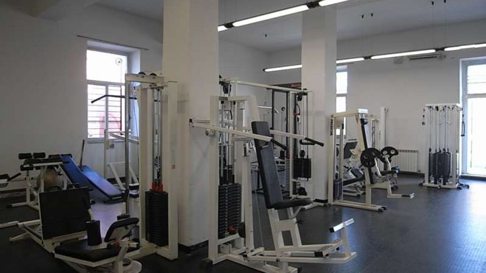 Interno di una palestra con pavimento scuro in gomma, grandi finestre e colonne bianche, illuminata da lunghe lampade a soffitto. Sono presenti varie macchine per allenamento con pesi e panche distribuite nella sala.