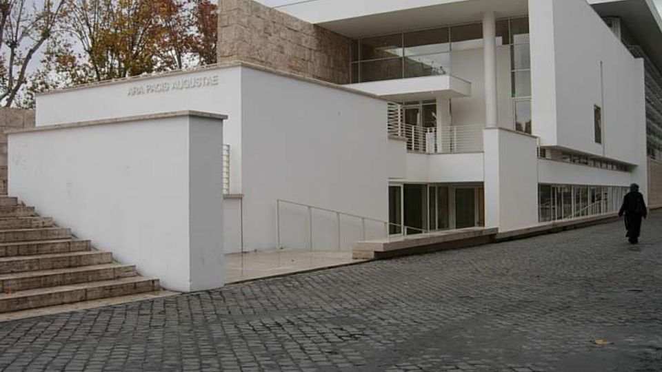 Edificio moderno bianco con volumi geometrici, grandi vetrate e pilastri, affiancato da un muro in pietra. Davanti si vede una strada in ciottoli con rampa e scala d’accesso, con alberi sullo sfondo.