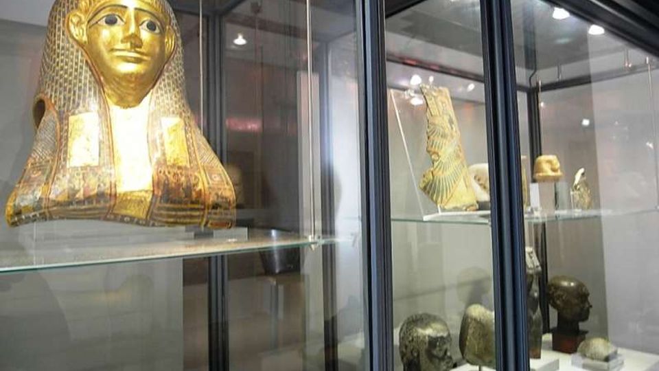 Vetrine museali in vetro e metallo che espongono reperti egizi, tra cui una maschera funeraria dorata e varie teste scultoree su piedistalli. L’ambiente interno è illuminato da faretti e presenta scaffali trasparenti con oggetti disposti su più livelli.