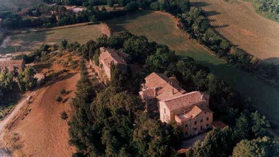 Vista aerea di una tenuta rurale con edifici in muratura dai tetti in coppi, circondati da alberi e campi coltivati. Intorno si estendono appezzamenti agricoli e strade di campagna che attraversano la campagna ondulata.