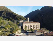 Veduta panoramica di un complesso religioso in pietra, con lunga facciata ad arcate e un campanile quadrato, situato in una valle tra colline boscose. Intorno si vedono strade e piccoli edifici con tetti in coppi, con montagne sullo sfondo.