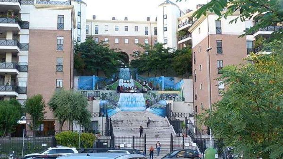 Grande scalinata urbana tra edifici residenziali in mattoni e intonaco, con terrazze laterali e rampe simmetriche. Al centro si vedono gradoni e strutture con teli blu simili a cascate, circondati da alberi e cancellate in ferro, con strada e auto in primo piano.
