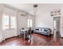 Soggiorno luminoso con pavimento in parquet scuro, pareti bianche e grandi finestre con tende chiare. Nella stanza ci sono un tavolo da pranzo con quattro sedie, un divano grigio e una lampada a sospensione, con passaggio verso altre stanze e mensole con vasi decorativi.