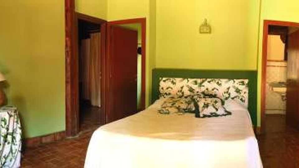 Camera da letto con pareti verde chiaro, letto matrimoniale al centro con testiera decorata, pavimento in cotto e mobili in legno. A sinistra c’è una finestra con tende a motivo floreale e una sedia in vimini, mentre a destra un comò con lampada e un’apertura verso un bagno.