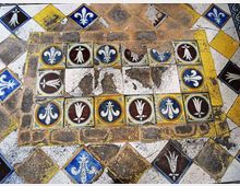 Pavimento decorato a mosaico con un riquadro centrale bordato da piastrelle colorate raffiguranti gigli stilizzati in blu, bianco e marrone. Intorno si vede una cornice di tessere romboidali e rettangolari in giallo, blu, nero e bianco, con segni di usura e lacune.