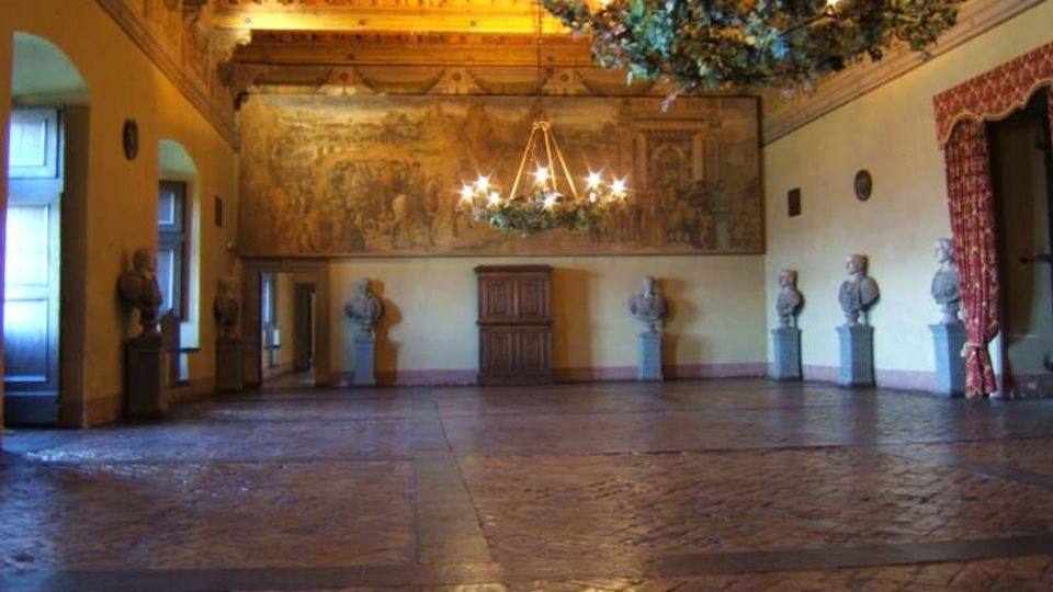 Ampia sala storica con pavimento in pietra e soffitto a travi lignee decorato, illuminata da grandi lampadari sospesi. Lungo le pareti sono disposti busti su piedistalli e un grande affresco, con finestre e porte laterali incorniciate da tendaggi.