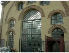 Facciata di un edificio in intonaco chiaro con grandi finestre ad arco e porta centrale vetrata a griglia, incorniciate da archi in mattoni. In primo piano si vedono una recinzione metallica e materiali da cantiere accatastati.