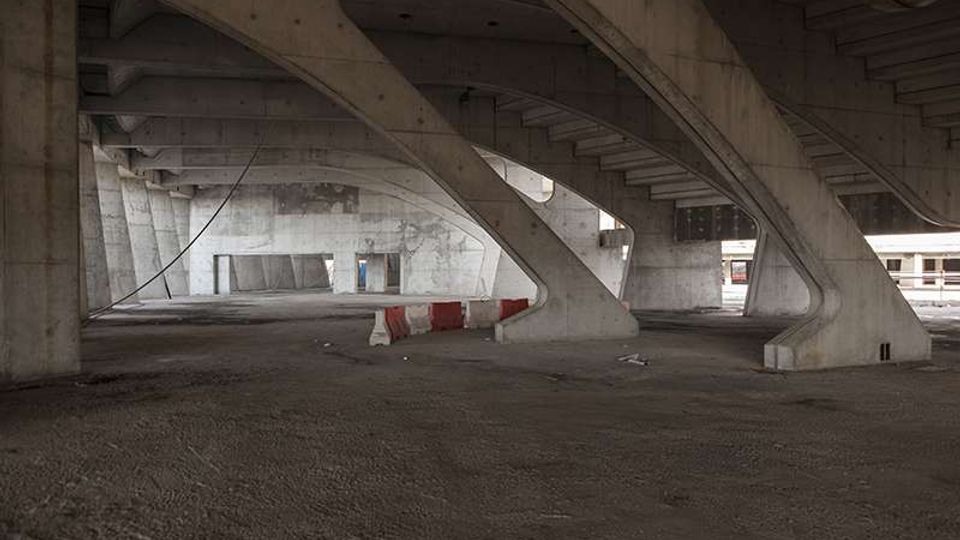 Spazio aperto sotto un cavalcavia in cemento armato, con grandi pilastri inclinati e travi a vista che sostengono la struttura. Pavimentazione in asfalto scuro con barriere stradali bianche e rosse e pareti di cemento sullo sfondo.