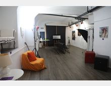 Sala espositiva con pareti bianche e pavimento in legno, con quadri appesi e file di sedie nere rivolte verso un fondale nero con cavalletto. Sono presenti luci da studio e un grande ombrello fotografico, oltre a una poltrona con coperta arancione e piccoli tavolini.