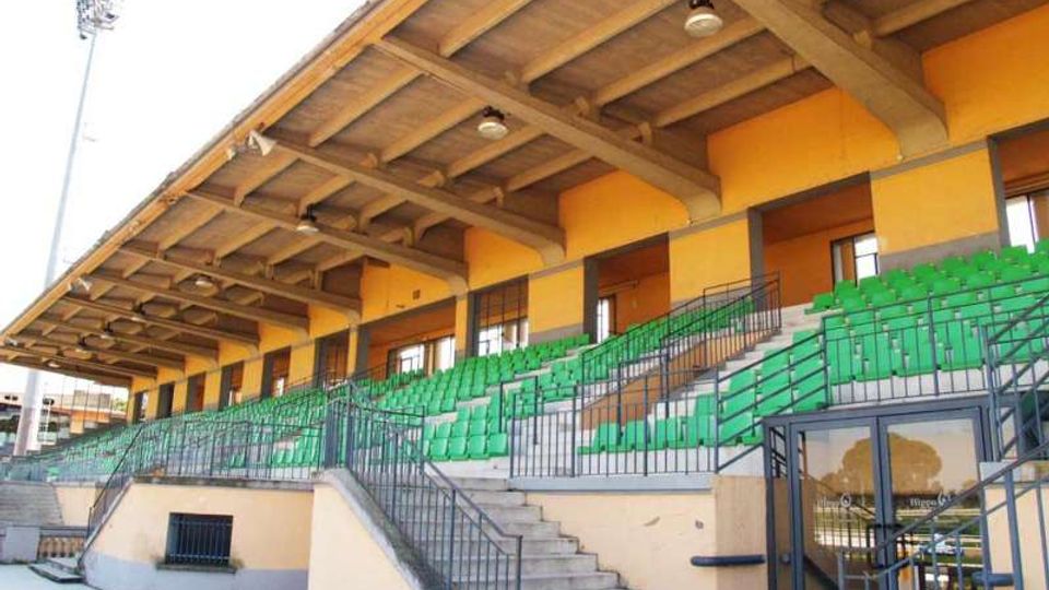 Tribuna coperta di uno stadio con struttura in cemento e travi a vista, file di sedili verdi disposte su gradinate. Scale e corrimano metallici conducono ai vari settori, con pareti giallo-arancio e ingresso vetrato alla base.