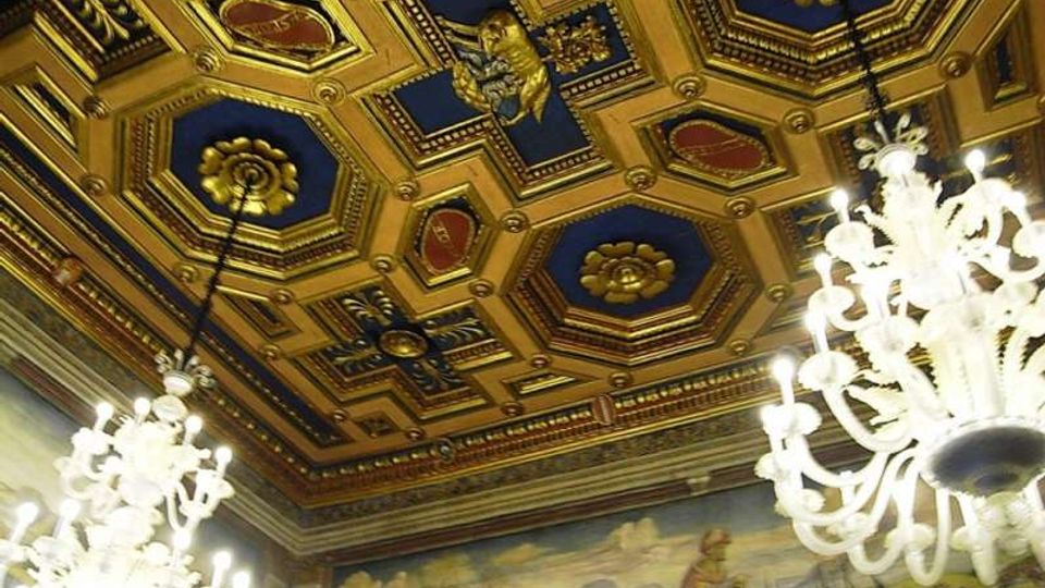 Soffitto interno di una sala storica con cassettonato dorato a riquadri geometrici blu e decorazioni in rilievo, con grandi lampadari in vetro sospesi. Sulle pareti corre un fregio dipinto con scene paesaggistiche e figure, sotto una cornice ornamentale.