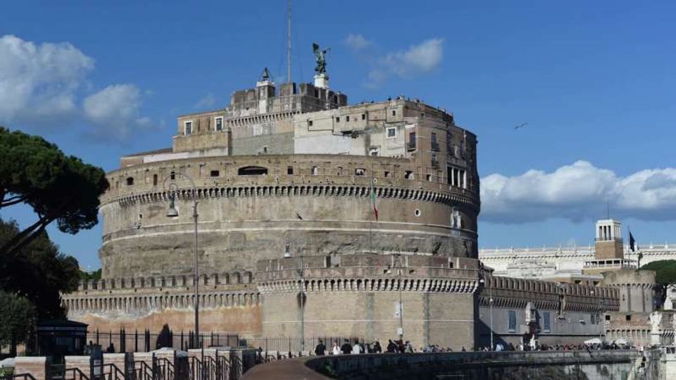Grande fortezza cilindrica in muratura con bastioni e merlature, affacciata sul lungofiume con una strada e recinzioni in primo piano. Sullo sfondo si vedono edifici adiacenti e un cielo azzurro con nuvole.