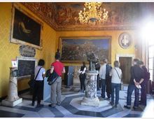 Sala museale con pareti giallo ocra decorate, grandi dipinti incorniciati e fregi affrescati vicino al soffitto, illuminata da un lampadario centrale. Al centro della stanza c’è una scultura su piedistallo in marmo e il pavimento è a motivi geometrici in pietra.