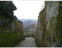 Stretto camminamento in pietra in discesa tra alte mura rocciose ricoperte di muschio e vegetazione, con una ringhiera in alto a destra. Sullo sfondo si apre una vista su colline e valli montuose.