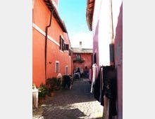 Vicolo stretto con pavimentazione in ciottoli tra edifici intonacati color arancio e rosa, con tetti in coppi e finestre con persiane. Lungo i muri si vedono vasi con piante e stendibiancheria con panni appesi, con vista su una piccola corte in fondo.