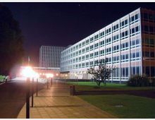 Veduta notturna di un complesso di edifici moderni a più piani con facciate a griglia di finestre illuminate. In primo piano un ampio vialetto pavimentato con lampioni, affiancato da prati e alcuni alberi.
