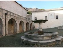 Cortile lastricato con una grande fontana in pietra al centro, composta da una vasca circolare e una coppa superiore su piedistallo. Sullo sfondo si vedono un porticato in pietra con archi e un edificio intonacato chiaro con finestre e una meridiana, con colline rocciose sullo sfondo.