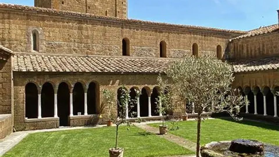 Chiostro in pietra di un complesso monastico con porticato ad archi su colonne, prato centrale e una fontana circolare in primo piano. Sullo sfondo si vede una torre campanaria rettangolare in muratura sotto un cielo sereno.