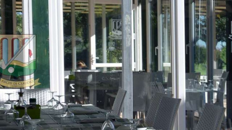 Terrazza coperta di un ristorante con tavoli apparecchiati e sedie intrecciate, affacciata su grandi vetrate con porte scorrevoli. Attraverso i vetri si vede un’area verde esterna con alberi e prato.