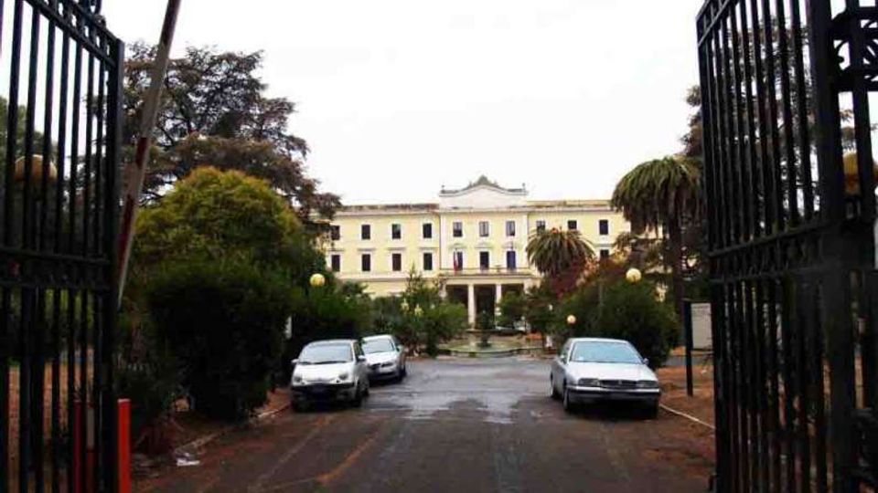 Ingresso con cancelli in ferro battuto aperti su un viale asfaltato che conduce a un grande edificio neoclassico a più piani, con facciata chiara e portico centrale. Ai lati del viale ci sono alberi e siepi, lampioni e alcune auto parcheggiate.