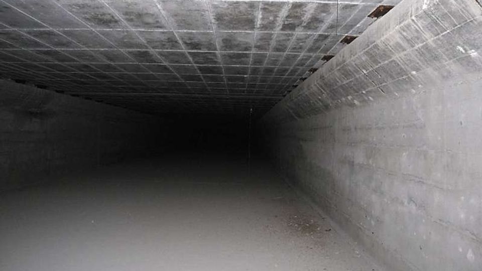 Interno di un lungo tunnel sotterraneo in cemento, con soffitto a griglia e pareti lisce che convergono verso un punto oscuro in lontananza. Il pavimento è spoglio e leggermente polveroso, illuminato debolmente.