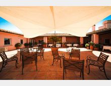 Cortile interno con pavimentazione in terracotta, sedute in legno e metallo disposte al centro e un grande telo bianco teso come copertura ombreggiante. Intorno si vedono edifici bassi color rosa con tetti in coppi e piante in vaso lungo il perimetro.