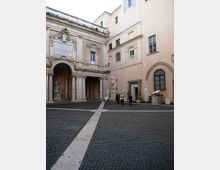Cortile interno in pietra con pavimentazione a sampietrini, circondato da edifici storici in stucco chiaro. A sinistra si vede un portico con archi e colonne classiche con iscrizione monumentale, mentre lungo le pareti sono esposte grandi sculture e frammenti in marmo.