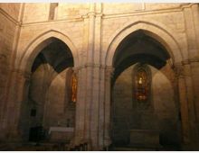 Interno di una chiesa in pietra con grandi archi a sesto acuto e pilastri centrali che sostengono volte ogivali. Sullo sfondo si vedono finestre strette con vetrate colorate e un piccolo altare laterale.