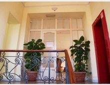 Interno di un edificio con pianerottolo e ringhiera in ferro battuto, pareti chiare e porta vetrata con finestre superiori. Ai lati della porta ci sono due grandi piante in vaso e sulla destra un vano o porta aperta con cornice rossa.