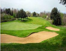 Campo da golf con ampio green e bandierina, circondato da fairway ondulato e alberi sullo sfondo. In primo piano si vedono due bunker di sabbia con bordi curvi lungo il lato destro del green.